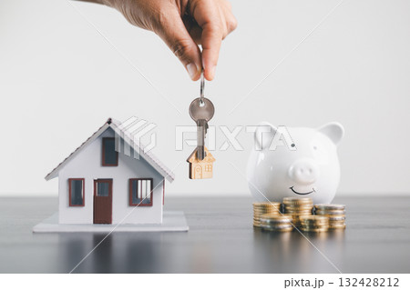 Hand holding house key above model home with piggy bank and stacked coins, symbolizing mortgage loan, real estate investment, savings, finance, property ownership and wealth management concept. Hand holding house key above model home with piggy bank and stacked coins, symbolizing mortgage loan, real estate investment, savings, finance, property ownership and wealth management concept. 132428212