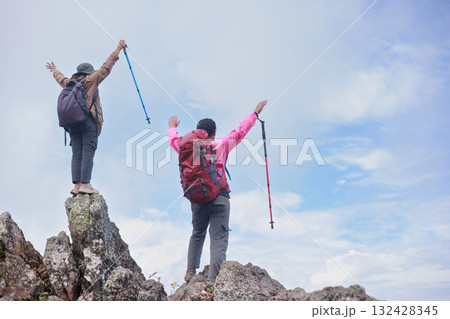 Two hikers with backpacks raising arms and trekking poles on rocky mountain peak, symbol of adventure, achievement, teamwork, freedom, success, exploration and outdoor lifestyle inspiration. 132428345