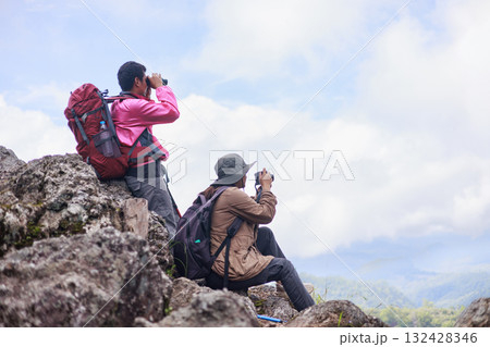 Two hikers with backpacks sitting on rocky mountain peak under blue sky, symbol of friendship, teamwork, freedom, inspiration, exploration, outdoor lifestyle and travel journey in nature. 132428346