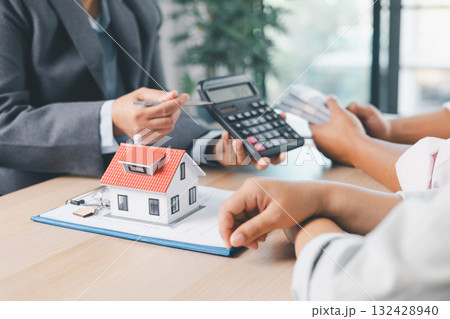 Close up of real estate agent handing house model to client with calculator and contract paperwork on desk, concept of mortgage loan, home ownership, property investment and housing agreement. 132428940