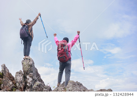 Two hikers with backpacks raising arms and trekking poles on rocky mountain peak, symbol of adventure, achievement, teamwork, freedom, success, exploration and outdoor lifestyle inspiration. Two hikers with backpacks raising arms and trekking poles on rocky mountain peak, symbol of adventure, achievement, teamwork, freedom, success, exploration and outdoor lifestyle inspiration. 132428944