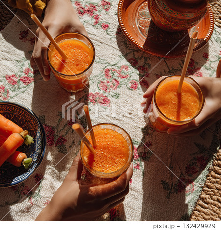Fresh carrot juice in three glasses with straws on floral tablecloth, hands holding glasses, rustic bowl of carrots and pottery plate creating warm cozy brunch scene Fresh carrot juice in three glasses with straws on floral tablecloth, hands holding glasses, rustic bowl of carrots and pottery plate creating warm cozy brunch scene 132429929