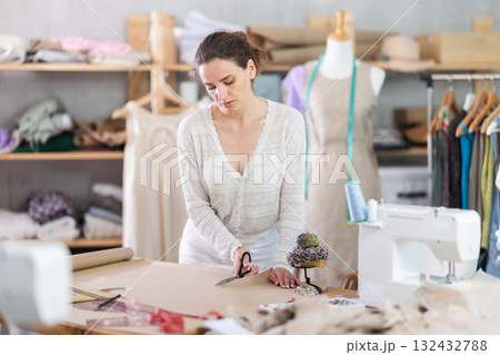 Female tailor cutting paper with scissors, cutting out the outline of pattern for a dress Female tailor cutting paper with scissors, cutting out the outline of pattern for a dress 132432788