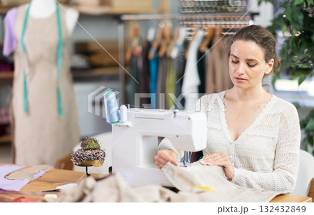 Working in sewing workshop - female dressmaker working at sewing machine making stitches on fabric Working in sewing workshop - female dressmaker working at sewing machine making stitches on fabric 132432849