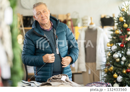Mature man trying on a down jacket against the background of a Christmas tree 132433560
