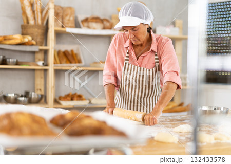 Skilled mature female baker rolling out dough in bakery 132433578