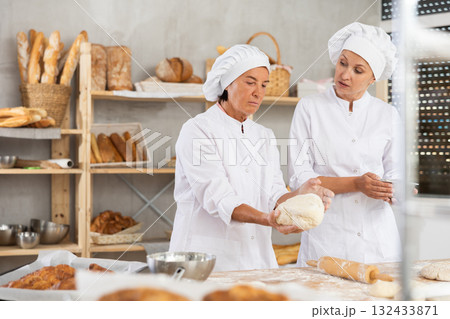 Elderly female baker giving advice to young assistant in kitchen Elderly female baker giving advice to young assistant in kitchen 132433871
