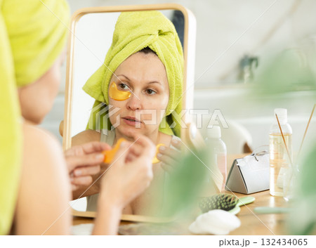 Middle-aged woman applying eye patch mask on her face sitting in front of the mirror 132434065