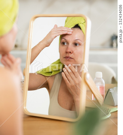Woman in towel turban on head examining facial skin in mirror 132434086