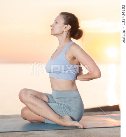 Sporty woman exercising yoga making Gomukhasana Cow Face Pose on seashore at sunrise 132434102
