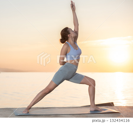 Active experienced woman teacher performs yoga asana on mat on seashore at sunset. Concept of healthy lifestyle 132434116