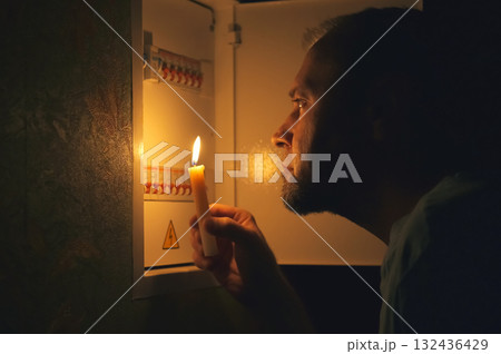 A man with a candle in the dark examines the electrical switchboard at home during a power outage. 132436429