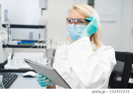 Blonde woman scientist wearing protective mask and lab coat, working on digital tablet in laboratory. Blonde woman scientist wearing protective mask and lab coat, working on digital tablet in laboratory. 132437231