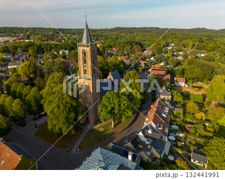 Aerial view of town with central Gothic style church, pointed steeple, red and gray rooftops, tree lined streets, and parked cars blending history with modern life. Aerial view of town with central Gothic style church, pointed steeple, red and gray rooftops, tree lined streets, and parked cars blending history with modern life. 132441991