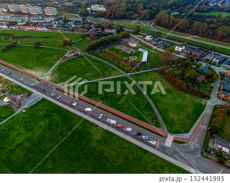 Aerial view of coastal urban recreational zone road with vehicles, red bike path, circular lookout, geometric green fields, tennis courts, and dense residential blocks framed by forest and shoreline. 132441993