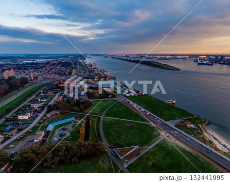 Aerial view of river port at sunset cargo ships, cranes, and industrial docks line both banks, with dense urban grid and green fields framing the waterway in a dynamic commercial landscape. 132441995