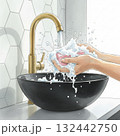 Hands are washing a bar of pink soap under running water. The water splashes over a black ceramic basin. The faucet is a modern, golden design 132442750