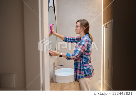 Woman wearing glasses cleaning round mirror with pink cloth near sink in modern bathroom. Concept of home cleaning, hygiene, daily routine and household chores 132442821