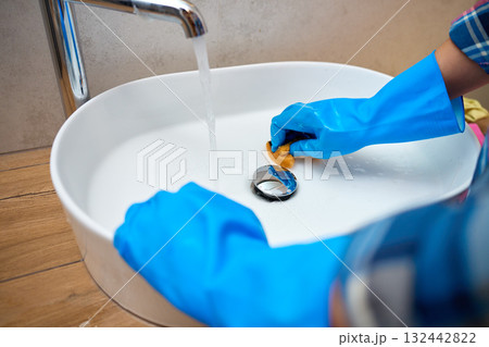 Woman wearing blue gloves cleaning cleaning white ceramic sink with sponge and soap foam in bathroom. Concept of hygiene, sanitation, household chores and daily home maintenance 132442822