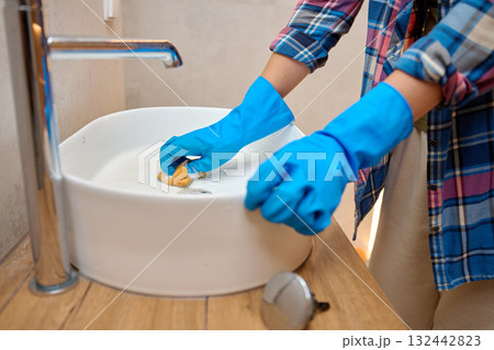 Woman wearing blue gloves cleaning cleaning white ceramic sink with sponge and soap foam in bathroom. Concept of hygiene, sanitation, household chores and daily home maintenance Woman wearing blue gloves cleaning cleaning white ceramic sink with sponge and soap foam in bathroom. Concept of hygiene, sanitation, household chores and daily home maintenance 132442823