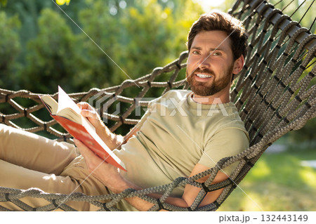 Caucasian bearded young man lying in hammock with a book in hands Caucasian bearded young man lying in hammock with a book in hands 132443149