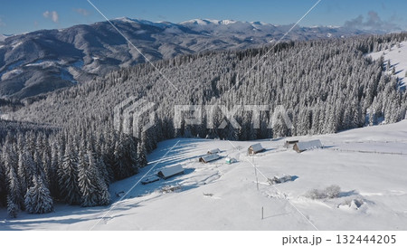 Breathtaking aerial drone revealing serene winter beauty snow covered mountain peaks, dense white blanketed forest, cozy houses nestled among frosty trees. Winter wild nature travel background 132444205