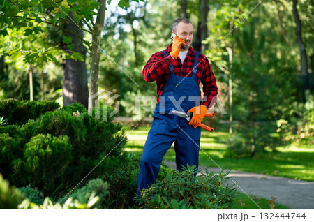 Gardener standing outdoors talking on mobile phone Gardener standing outdoors talking on mobile phone 132444744