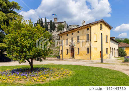 Sunny spring day at Brescia Castle park with blooming flowers yellow walls and flags on tower 132445780