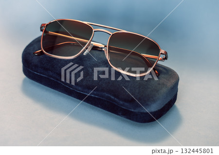 Modern sunglasses with metallic frame and tinted lenses rest on navy velvet case pastel backdrop Modern sunglasses with metallic frame and tinted lenses rest on navy velvet case pastel backdrop 132445801