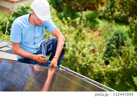 Worker building photovoltaic solar panel system on roof of house. Engineer in helmet looking on evenness of solar modules during installing outdoors. Concept of alternative and renewable energy. Worker building photovoltaic solar panel system on roof of house. Engineer in helmet looking on evenness of solar modules during installing outdoors. Concept of alternative and renewable energy. 132447895