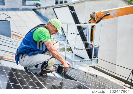 Worker building photovoltaic solar panel system on metal rooftop of house. Man technician installing solar module with help of electric drill outdoors. Alternative energy generation concept. 132447904