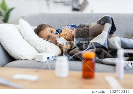 A young child lies on a couch, using a nebulizer to help with breathing while holding a teddy bear, with medication bottles in the foreground. A young child lies on a couch, using a nebulizer to help with breathing while holding a teddy bear, with medication bottles in the foreground. 132453313