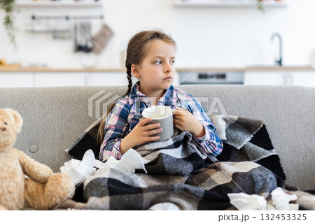A young girl, wrapped in a blanket, looks unwell while holding a mug. Tissues and a teddy bear sit beside her, suggesting she is recovering from an illness. 132453325