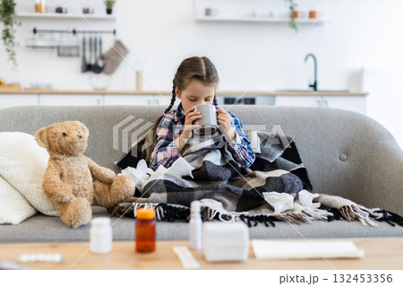 A young girl, wrapped in a blanket, sits on a couch and drinks from a mug, surrounded by medicine and tissues, looking unwell. A young girl, wrapped in a blanket, sits on a couch and drinks from a mug, surrounded by medicine and tissues, looking unwell. 132453356