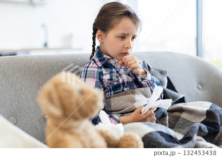 A young girl, wrapped in a blanket, coughs while holding a tissue. A teddy bear sits in the foreground, suggesting comfort. A young girl, wrapped in a blanket, coughs while holding a tissue. A teddy bear sits in the foreground, suggesting comfort. 132453438