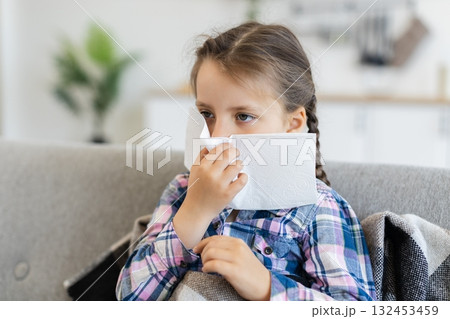 A young girl with braids is sitting on a couch, holding a tissue to her nose, possibly suffering from a cold or allergies. A young girl with braids is sitting on a couch, holding a tissue to her nose, possibly suffering from a cold or allergies. 132453459