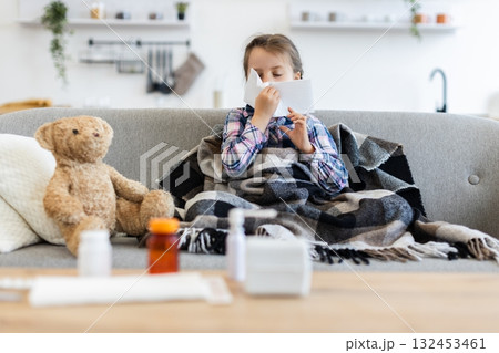A young girl, wrapped in a blanket, sits on a couch, blowing her nose with a tissue. A teddy bear sits beside her, and medicine is on the table. A young girl, wrapped in a blanket, sits on a couch, blowing her nose with a tissue. A teddy bear sits beside her, and medicine is on the table. 132453461