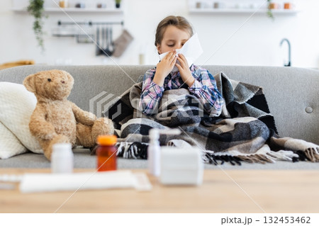 A young girl, wrapped in a blanket, is blowing her nose with a tissue while sitting on a couch. A teddy bear and medicine are nearby. A young girl, wrapped in a blanket, is blowing her nose with a tissue while sitting on a couch. A teddy bear and medicine are nearby. 132453462