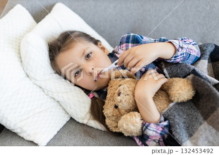 A young girl, wrapped in a blanket, lies on a sofa while taking her temperature with a thermometer. She is holding a teddy bear for comfort. 132453492