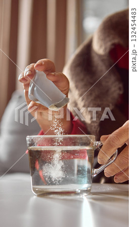 Elderly hand pouring powdered supplement into glass cup of hot water Elderly hand pouring powdered supplement into glass cup of hot water 132454483