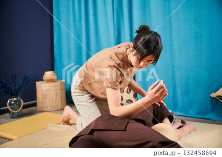 Masseuse making thai yoga massage. Woman therapist doing traditional massage treatment for female patient using elbow. Client lying on mat, meditating, enjoying hand massage in calm atmosphere. 132458694
