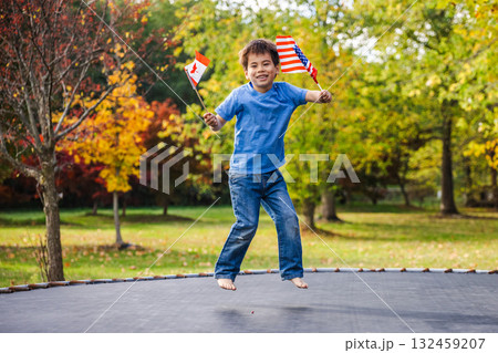 Cheerful boy jumps barefoot on a trampoline holding USA and Canada flags in a colorful autumn park Cheerful boy jumps barefoot on a trampoline holding USA and Canada flags in a colorful autumn park 132459207
