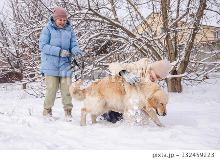 Elderly woman and her adult daughter playing with a Golden Retriever in the snow. The daughter pets the cheerful dog while the mother holds the leash and smiles, enjoying a joyful winter moment Elderly woman and her adult daughter playing with a Golden Retriever in the snow. The daughter pets the cheerful dog while the mother holds the leash and smiles, enjoying a joyful winter moment 132459223