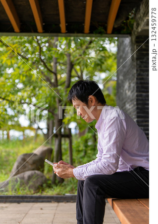 公園のベンチで通知を確認するビジネス男性、緑に囲まれたベンチでスマホに集中する男性 132459778
