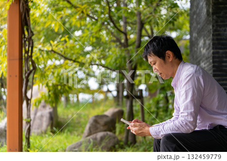 公園のベンチで通知を確認するビジネス男性、緑に囲まれたベンチでスマホに集中する男性 132459779