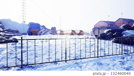Snow blankets the ground at a secluded research station, with colorful structures peeking through the icy landscape. Tall rocks stand around, bathed in a serene winter light. 132460040