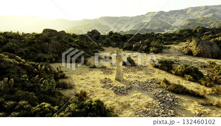 A towering stone statue stands alone in a vast, rugged terrain surrounded by rocky hills and dense greenery. The sun casts long shadows in the tranquil atmosphere. 132460102