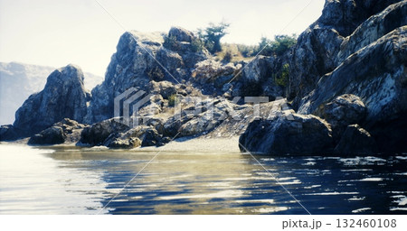 Rugged rocks meet serene waters on a tranquil coastline. Sunlight illuminates the area, highlighting the natural beauty and distant hills. A peaceful setting invites exploration. 132460108
