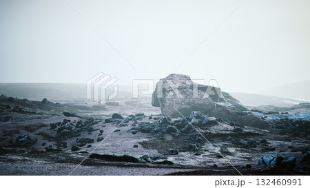 A vast, ethereal landscape unfolds with a massive boulder resting amid a barren, rocky expanse. The scene is enveloped in a cloak of mist, creating an otherworldly atmosphere. 132460991