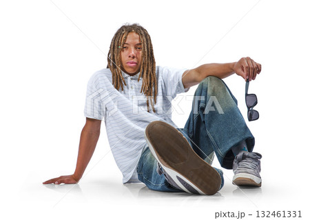 Young man with dreadlocks sitting on floor holding sunglasses Young man with dreadlocks sitting on floor holding sunglasses 132461331